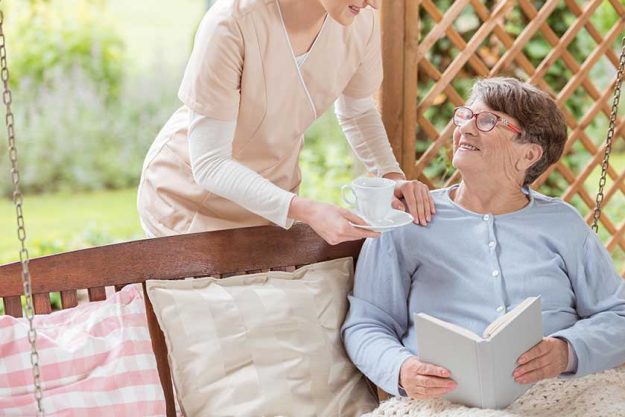 Nurse giving tea to happy elderly woman reading book