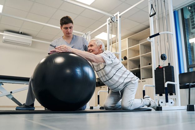 Pensioner standing on knees while putting arms on exercise ball Pensioner standing on knees while putting arms on exercise ball