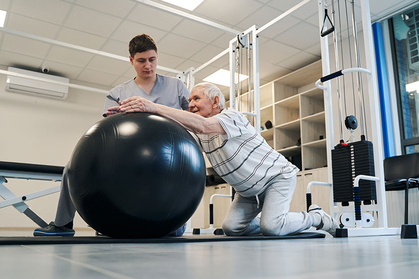 Pensioner standing on knees while putting arms on exercise ball
