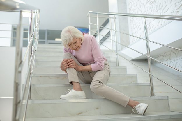 Senior woman in pink shirt sitting on the stairs with a damaged knee Senior woman in pink shirt sitting on the stairs with a damaged knee