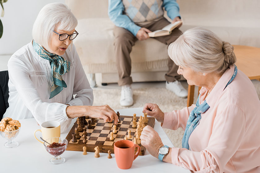 Senior women playing chess and drinking coffee while man reading book on sofa Senior women playing chess and drinking coffee while man reading book on sofa